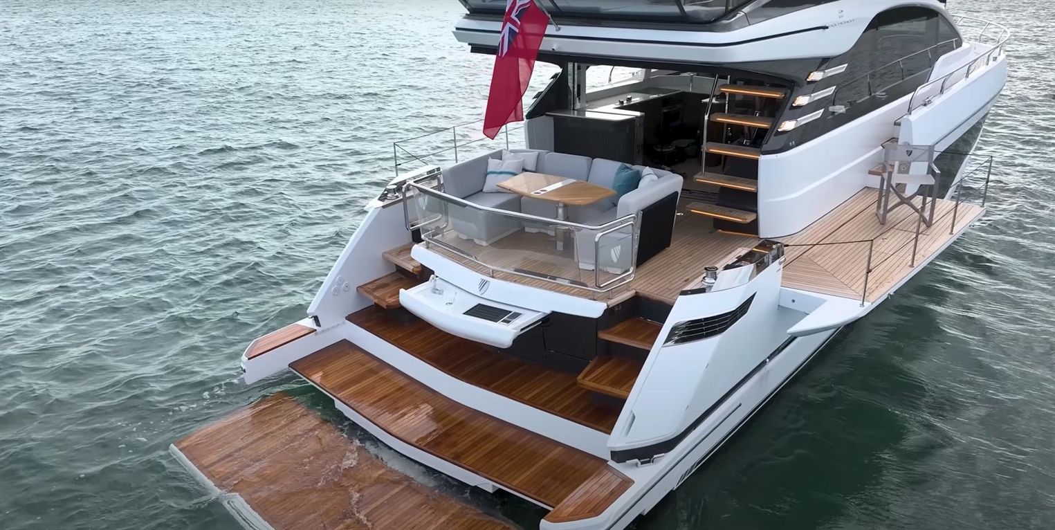 A luxury yacht with a spacious deck, large lounge area, and wooden steps on tranquil water, featuring a flag at the rear.